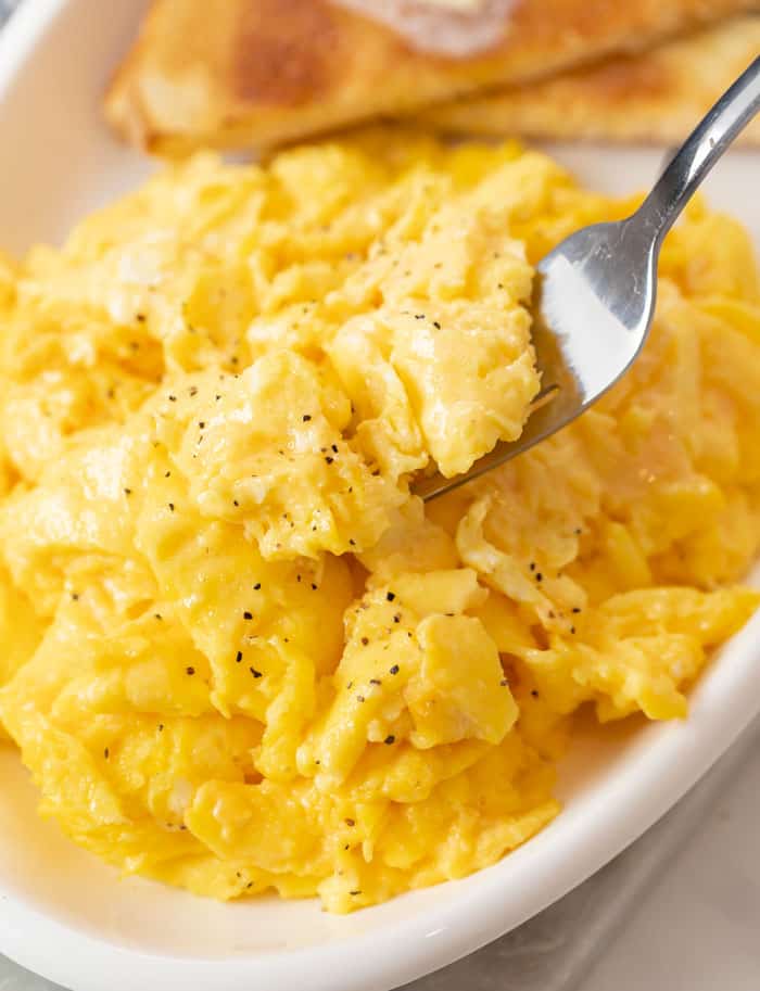 Une assiette d'œufs brouillés moelleux avec une fourchette les ramassant et des toasts en arrière-plan.