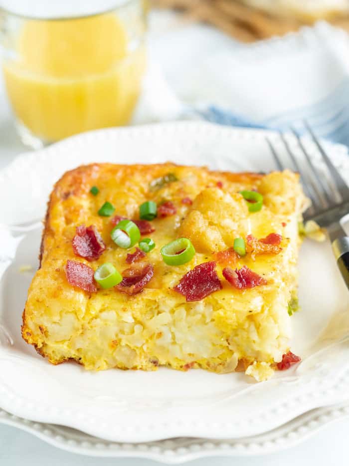 Une tranche de casserole de petit-déjeuner tater tot sur une plaque blanche avec une fourchette et du jus d'orange en arrière-plan.