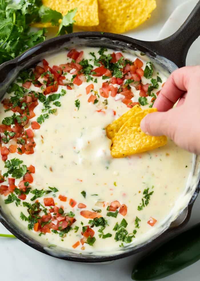 Une main trempant une tortilla chips dans une poêle de trempette au queso blanc garnie de Pico De Gallo et de coriandre